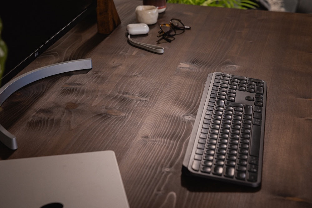 Close up of a dark stained wooden desk with a keyboard, monitor stand and small accessories arranged neatly on the surface.