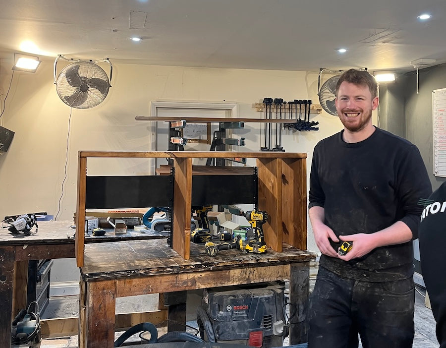 A smiling furniture maker in a workshop standing beside a handmade wooden desk in progress, surrounded by tools and woodworking equipment.