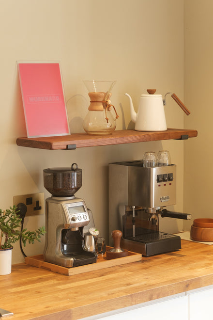 Kitchen coffee station with espresso machine, grinder and Chemex on a wooden worktop, with a matching wooden shelf above holding coffee accessories.