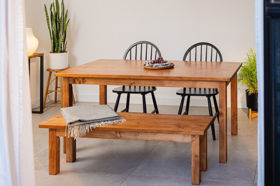 Handmade solid pine dining table with matching wooden bench, crafted from sustainably sourced softwood by Pastos & Co, shown in a minimalist dining space with black chairs and indoor plants.