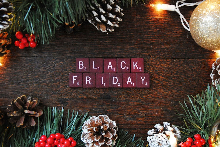 Black Friday text spelled with Scrabble tiles surrounded by pinecones, berries and Christmas lights on a wooden surface.