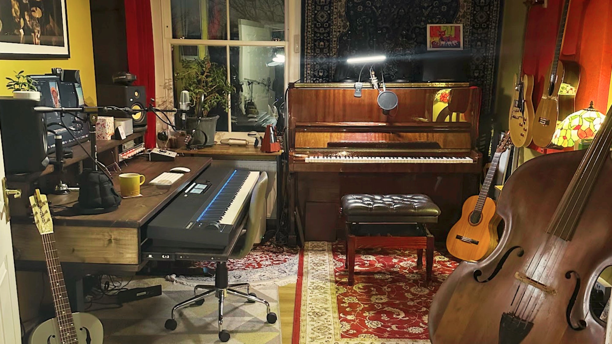 Home music studio setup with wooden desk, MIDI keyboard, studio monitors, upright piano, guitars, and acoustic instruments in a warm, lived-in room