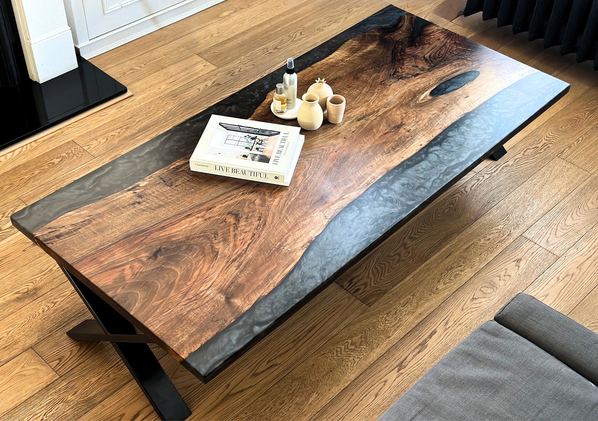 A rectangular lounge table with a walnut wood top and black resin detailing, set on steel legs, with a small object on top and a book beside it on a wooden floor.