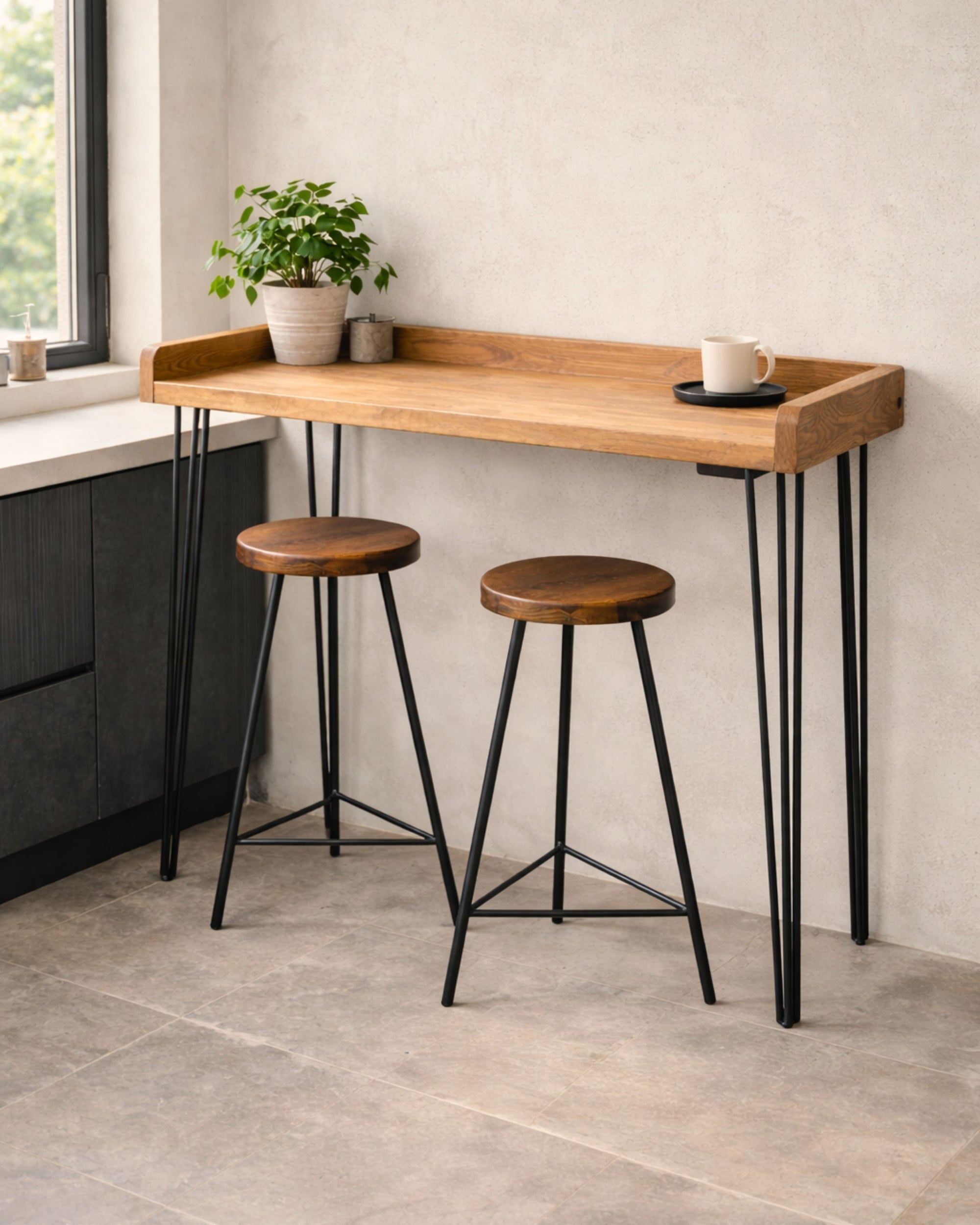 Handmade solid wood breakfast bar table with black hairpin legs and two stools beside a kitchen window, styled with a plant and coffee cup.