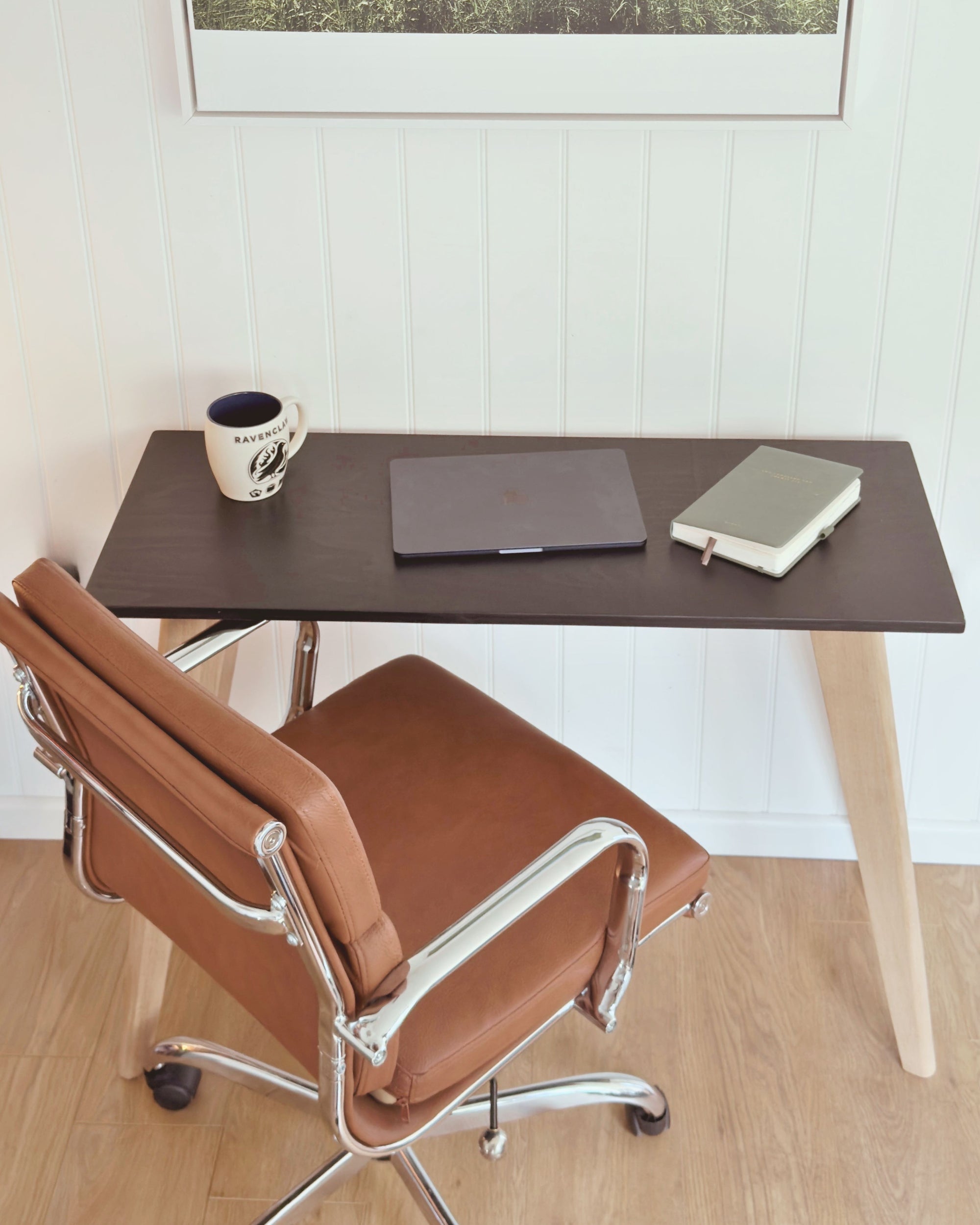 Scandi style minimalist desk with a black birch plywood top and natural light oak legs, set against a white panelled wall. A laptop, notebook and mug sit on the surface, with a brown leather office chair beside it.