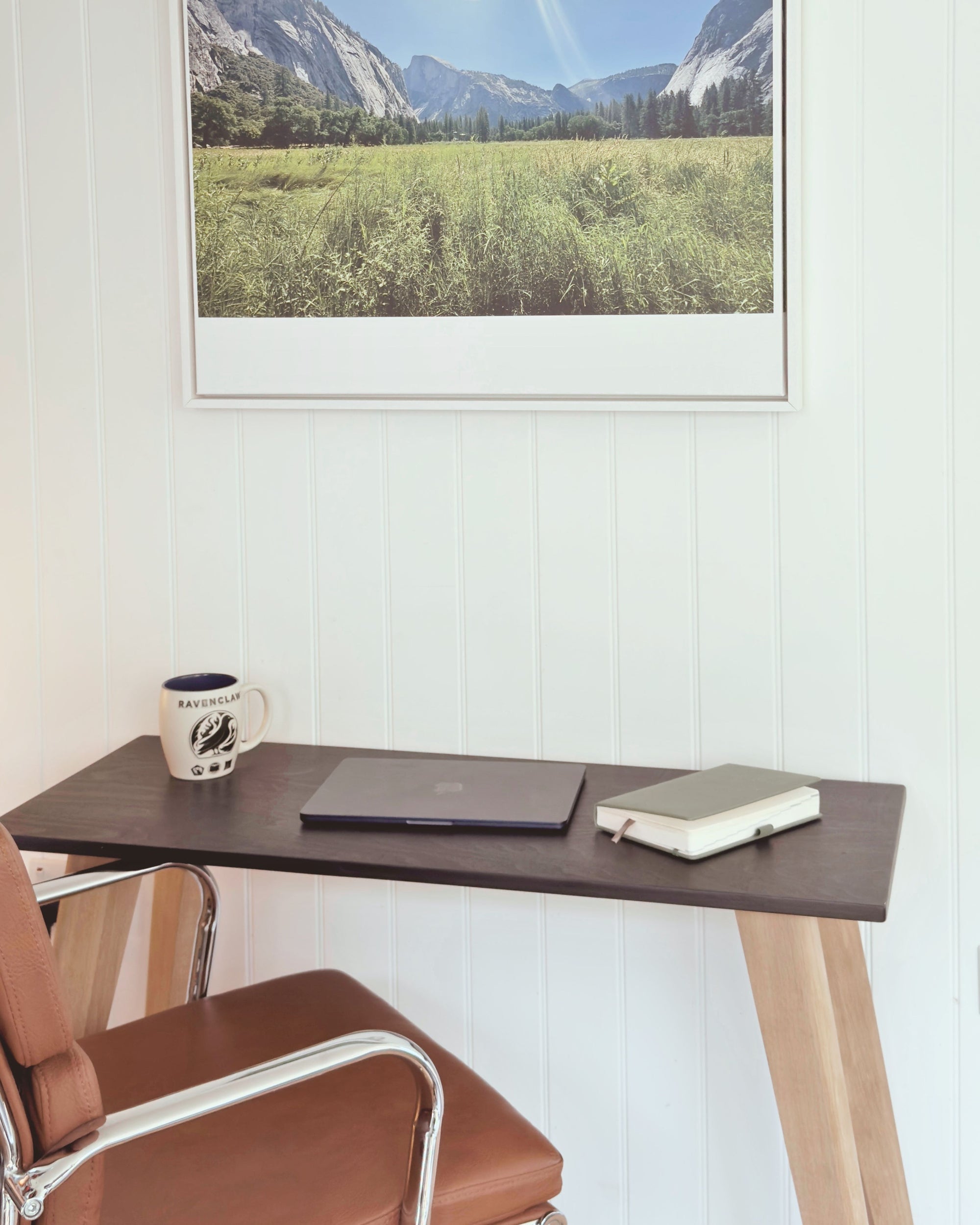 Scandi style minimalist desk with a black birch plywood top and natural light oak legs, set against a white panelled wall. A laptop, notebook and mug sit on the surface, with a brown leather office chair beside it.