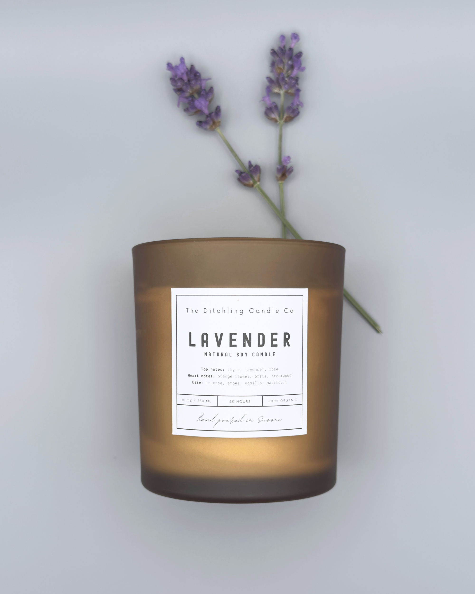 Natural soy, lavender scented candle in a 10oz amber glass, with white label and matt grey background and two lavender sprigs next to it. Top notes of thyme, lavender and rose. Heart notes of orange, flower, orris and cedarwood. Base notes of incense, amber, vanilla and patchouli. Finished with a wooden wick.