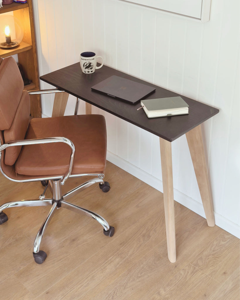 Scandi style minimalist desk with a black birch plywood top and natural light oak legs, set against a white panelled wall. A laptop, notebook and mug sit on the surface, with a brown leather office chair beside it.