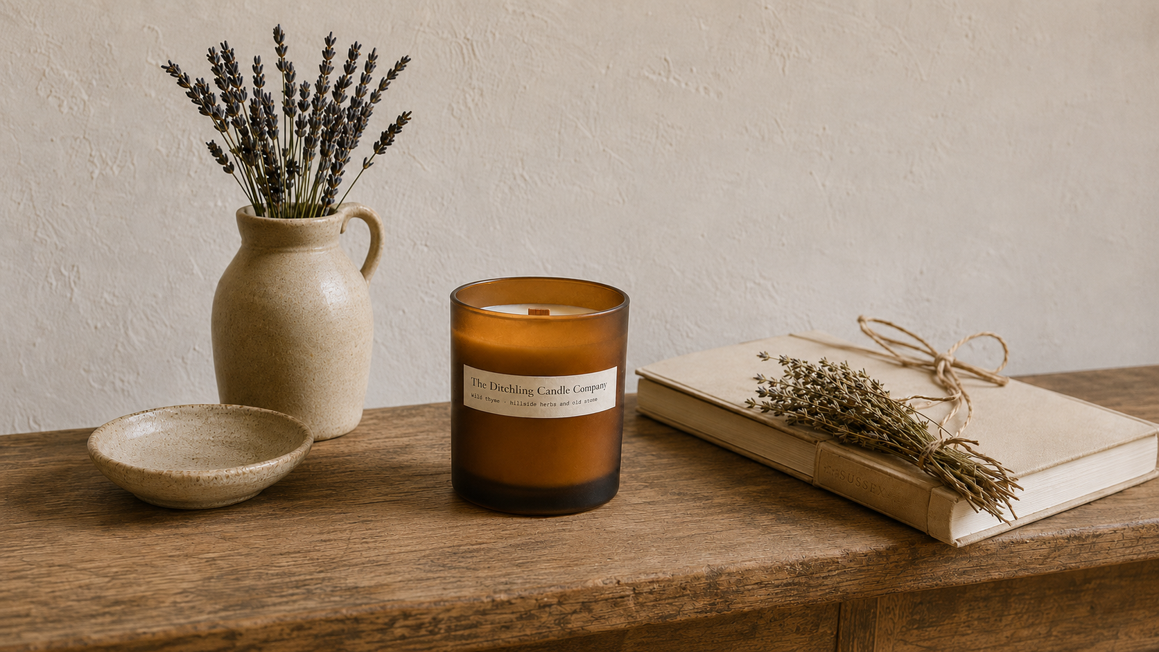 An amber glass jar scented soy candle with wooden wick and with a parchment coloured label reading 'The Ditching Candle Company' and with scent name 'wild thyme' with notes of 'hillside herbs and old stone' sits on a rustic wooden table, styled with dried lavender in a ceramic pitcher and a book in the background with a bunch of wild thyme on it.